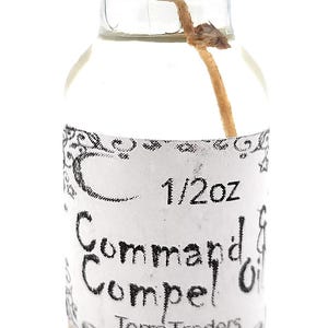 Peut inclure: Petite bouteille en verre avec un bouchon noir, contenant un liquide clair. L'étiquette indique "Command Compel Oil" et "1/2oz" avec le logo Terra Traders. Une fibre naturelle est visible à l'intérieur.