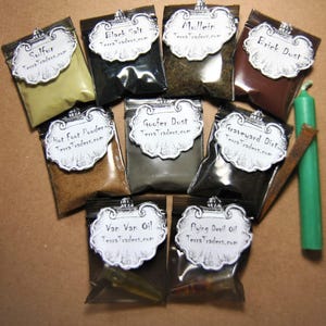 May include: An assortment of small, sealed bags containing various powders and oils, labeled with ornate tags. The bags are filled with substances like sulfur, black salt, and brick dust. A green candle and a piece of wood are also present. The tags read "TerraTraders.com".