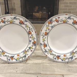 May include: Two round white ceramic plates featuring a decorative border pattern of thin brown branches with small orange, yellow, and blue flowers. The outer edge of each plate is finished with a thin black and white geometric chain-link design.