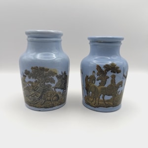 Peut inclure: Deux pots en céramique bleu clair ornés d'illustrations en relief marron représentant des personnages à cheval et des arbres. Chaque pot possède un col large et une finition lisse et brillante, illustrant une scène de chasse sur le pourtour.