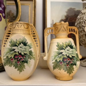 Könnte beinhalten: Zwei cremefarbene Keramikvasen mit einem bemalten Blumenmuster aus grünen Blättern und kleinen rosa Beeren. Die linke Vase hat eine ovale Form mit seitlichen Gittergriffen, während die rechte Vase einen schmalen Hals und zwei Bogengriffe hat.