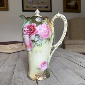 May include: A porcelain pitcher featuring a hand-painted floral design with large pink and red roses against a soft yellow and green background. The vessel has a scalloped rim, a curved handle, and a matching lid with a pointed finial top.