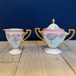 May include: A porcelain creamer and a lidded sugar bowl featuring a cream and pale pink color palette with hand-painted floral motifs in pink and blue. Both pieces have gold-painted handles, rims, and bases, with a hexagonal footed design.