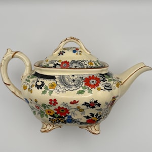 May include: A cream-colored ceramic teapot featuring a vibrant floral pattern in red, yellow, green, and black. The teapot has a curved handle, a pointed spout, a matching lid with a handle, and stands on three small, ornate feet.