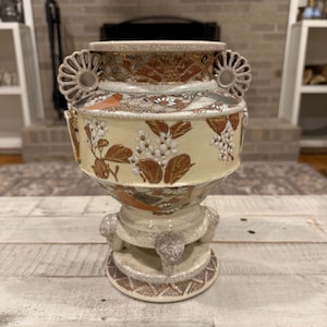 May include: A ceramic vase with a cream-colored base, featuring intricate gold, brown, and white floral patterns. The vessel has two decorative side handles, a wide rim, and a pedestal base supported by three small, textured, claw-like feet.