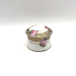 May include: A small, round porcelain trinket box with a white base and a lid. The exterior features hand-painted pink rose blossoms, a grey band, and intricate gold filigree detailing along the rim and sides. The lid has a white center.