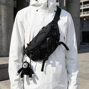 May include: Black fanny pack with multiple compartments and adjustable straps. A small stuffed monkey keychain hangs from the pack. The person is wearing a white hooded jacket.