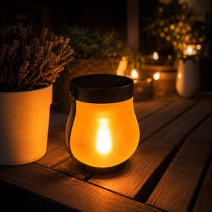 Könnte beinhalten: Eine schwarze und bernsteinfarbene Laterne mit Griff steht auf einem Holztisch. Die Laterne hat ein warmes, leuchtendes Licht im Inneren und schafft eine gemütliche Atmosphäre. Weitere Laternen und Pflanzen sind im Hintergrund zu sehen, was auf eine Außendekoration hindeutet.
