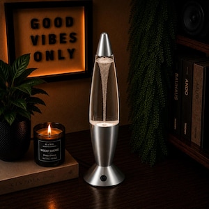 Peut inclure: Une lampe tornade argentée avec une chambre cylindrique en verre transparent contenant un vortex de bulles tourbillonnantes. La lampe présente une base en argent métallique et un capuchon pointu assorti, avec un petit bouton noir sur la base.