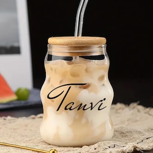 Könnte beinhalten: Ein durchsichtiges, dosenformiges Glas mit gewellter Silhouette, gefüllt mit einem Eiskaffee-Getränk. Das Glas ist mit dem Namen Tanvi in schwarzer Schreibschrift, einem hellen Bambusdeckel und einem durchsichtigen Glasstrohhalm versehen.