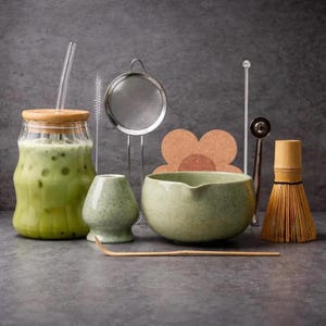May include: A matcha tea set including a glass jar with a bamboo lid and straw, a metal strainer, a light green ceramic bowl and whisk holder, a bamboo whisk, a bamboo scoop, a cork coaster, a cleaning brush, and a metal stirring tool.