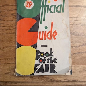 May include: A vintage paperback book titled Official Guide Book of the Fair with a white cover featuring large green, orange, and yellow circles. The text is printed in black, green, and orange, with a 25 cent price mark in a red circle.