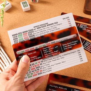 May include: A Mah Jongg score card featuring a white background with black, red, and green text, partially covered by a rectangular tortoiseshell-patterned plastic viewing guide. The card lists various hand combinations, point values, and game rules.