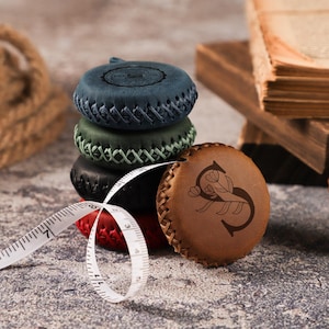 Pode incluir: Uma pilha de quatro fitas métricas redondas de couro nas cores azul, verde, preta e vermelha, com uma fita métrica de couro bege à frente com um design de letra S floral. Uma fita métrica branca com marcas pretas em cm está enrolada à frente.