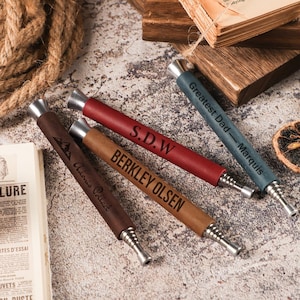 May include: Four cylindrical fire bellows tools with metallic tips and leather-like sleeves in dark brown, tan, red, and blue-grey. Each tool features custom laser-engraved text including names like Berkley Olsen, S.D.W, and Greatest Dad - Marquis.