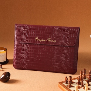 May include: A rectangular burgundy laptop sleeve with a textured crocodile skin pattern and a flap closure. The name Grayson Thomas is embossed in gold lettering on the front flap. The sleeve is positioned horizontally against a solid background.