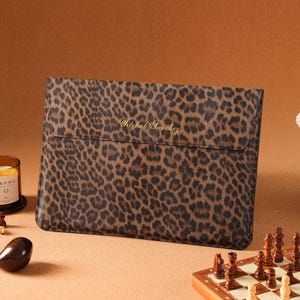 Könnte beinhalten: Eine rechteckige Laptoptasche mit einem braun-schwarzen Leopardenmuster. Die vordere Klappe ist mit dem goldfarbenen Schriftzug Yitzhak Sanchez in einer kursiven Schriftart personalisiert. Die Tasche hat ein schmales Profil und einen Umschlag.