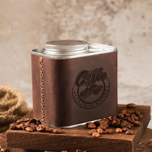 Pode incluir: Um recipiente metálico retangular envolto em couro castanho escuro com uma costura cruzada visível na lateral. O couro apresenta um logótipo circular com o texto Coffee e Coleman Joyner. Tem uma tampa prateada de rosca na parte superior.