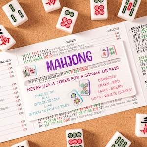 May include: A Mahjong score card featuring printed rules, hand combinations, and point values in purple, red, green, and black text, accompanied by scattered white Mahjong tiles with red and green character and suit markings.