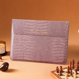 May include: A rectangular, mauve-coloured laptop sleeve featuring a textured crocodile skin pattern. The front flap displays the gold-embossed text "Karsten Schultz" centred horizontally. The sleeve has a slim, envelope-style design.