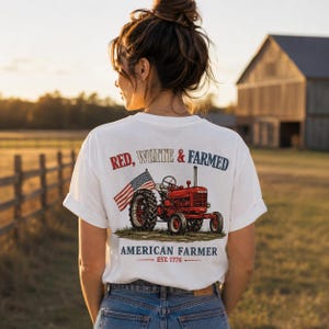 Könnte beinhalten: Weißes T-Shirt mit einem roten Traktorgrafik, amerikanischer Flagge und dem Text "RED, WHITE & FARMED" und "AMERICAN FARMER EST. 1776". Das T-Shirt wird von einer Person in ländlicher Umgebung getragen.