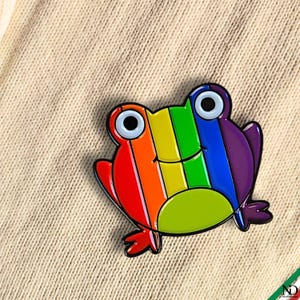 Puede incluir: Un pin de esmalte metálico con forma de rana y cuerpo a rayas de arcoíris que incluye secciones en rojo, naranja, amarillo, verde, azul y morado. La rana tiene dos ojos grandes blancos con pupilas negras y una línea negra como boca.