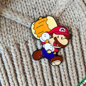 Puede incluir: Un pin de esmalte metálico que muestra un personaje de dibujos animados con una gorra roja con un símbolo M blanco, una camisa azul y zapatos marrones, sosteniendo un gran martillo amarillo con una cabeza de color crema claro.