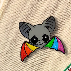 Op de afbeelding: Een metalen emaille speld in de vorm van een vleermuis met een grijze kop, grote zwarte ogen en kleine witte hoektanden. De vleermuis heeft uitgestrekte vleugels met een levendig regenboogverloop en een glinsterende afwerking.