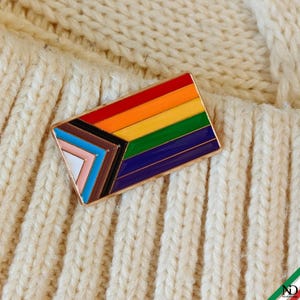 Może przedstawiać: Prostokątna emaliowana przypinka z wzorem flagi Progress Pride. Przypinka posiada poziome paski w kolorach czerwonym, pomarańczowym, żółtym, zielonym, niebieskim i fioletowym oraz klin po lewej stronie w kolorach czarnym, brązowym, jasnoniebieskim, różowym i białym.