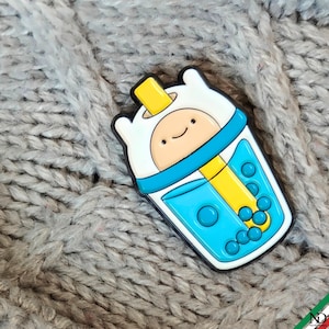 Puede incluir: Un pin de esmalte colorido con forma de taza de té de burbujas que presenta un personaje de dibujos animados con una capucha blanca, una base de líquido azul, una pajita amarilla y varias pequeñas perlas de boba azules dentro de la taza.