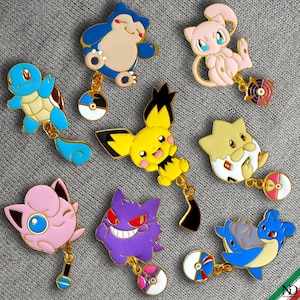 Può includere: Una collezione di spille smaltate colorate raffiguranti vari personaggi dei Pokémon. Ogni spilla ha un design dettagliato ed è attaccata a un piccolo ciondolo. Le spille sono poste su uno sfondo grigio.
