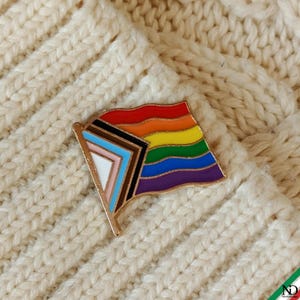 Könnte beinhalten: Eine Metall-Emaille-Anstecknadel in Form einer wehenden Flagge mit dem Progress-Pride-Design. Die Flagge zeigt horizontale Streifen in Rot, Orange, Gelb, Grün, Blau und Violett sowie einen Chevron auf der linken Seite in Schwarz, Braun, Hellblau, Rosa und Weiß.
