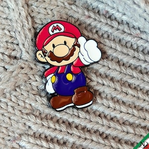Puede incluir: Un pin de esmalte metálico que muestra un personaje de dibujos animados con una gorra roja con una M blanca, bigote marrón, camisa roja, peto azul con botones amarillos, guantes blancos y zapatos marrones, en una pose de pie con una mano alzada.