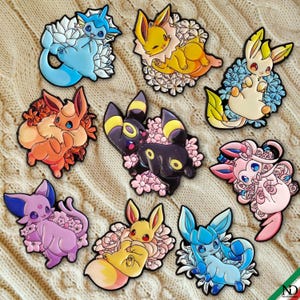 May include: A collection of nine enamel pins featuring various Eevee evolution characters, each surrounded by distinct floral arrangements. The pins display vibrant colors including blue, yellow, orange, black, pink, and purple, with detailed outlines.