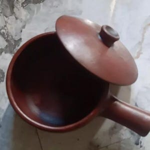 May include: A terracotta-coloured cooking pot with a lid. The pot has a handle and a round body. The lid is slightly ajar, revealing the dark interior. The pot is likely made of clay and is suitable for cooking.