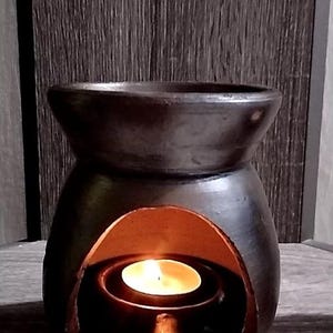 May include: A dark brown ceramic essential oil diffuser with a lit tea light candle inside. The diffuser has a bulbous body with a wide opening at the top and a curved opening in the front. The candle flame glows brightly.