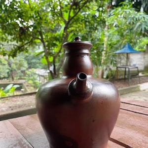 May include: A brown ceramic water jug with a spout and a lid. The jug is sitting on a wooden table. The background is blurred, showing green foliage and a glimpse of a blue structure.