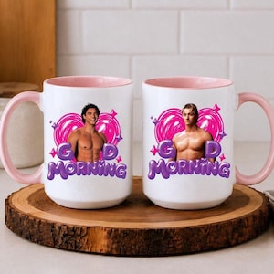 May include: Two white ceramic mugs with light pink interiors and handles. Each mug features a graphic of a shirtless person centred within a bright pink heart outline, accompanied by the purple text "GD MORNING" in a bold, rounded font.