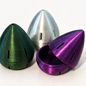 May include: Three metallic-looking, cone-shaped objects. One is green, one is silver, and one is purple and open, revealing a matching interior. Each has rectangular cutouts. The objects are on a white surface.