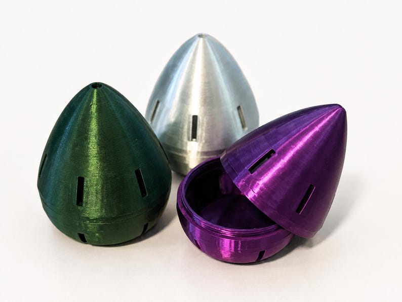 May include: Three metallic-looking, cone-shaped objects. One is green, one is silver, and one is purple and open, revealing a matching interior. Each has rectangular cutouts. The objects are on a white surface.