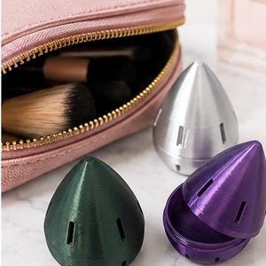 May include: A pink makeup bag with a gold zipper, makeup brushes, and three cone-shaped containers in green, silver, and purple. The purple container is open, revealing its interior.