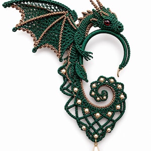 Könnte beinhalten: Ein dunkelgrüner und bronzefarbener Ohrschmuck in Drachenform aus filigraner Fadenarbeit. Das Design zeigt einen detaillierten Drachenkopf mit rotem Auge, ausgebreiteten Flügeln und einem geringelten Schwanz, verziert mit cremefarbenen Perlen.