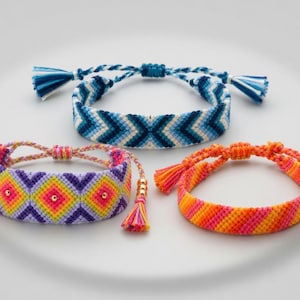 Pode incluir: Três pulseiras da amizade tecidas com padrões intrincados. Uma apresenta um design de chevron azul, outra um motivo de diamante roxo e amarelo com contas douradas, e a terceira mostra listras diagonais laranja e rosa com borlas.