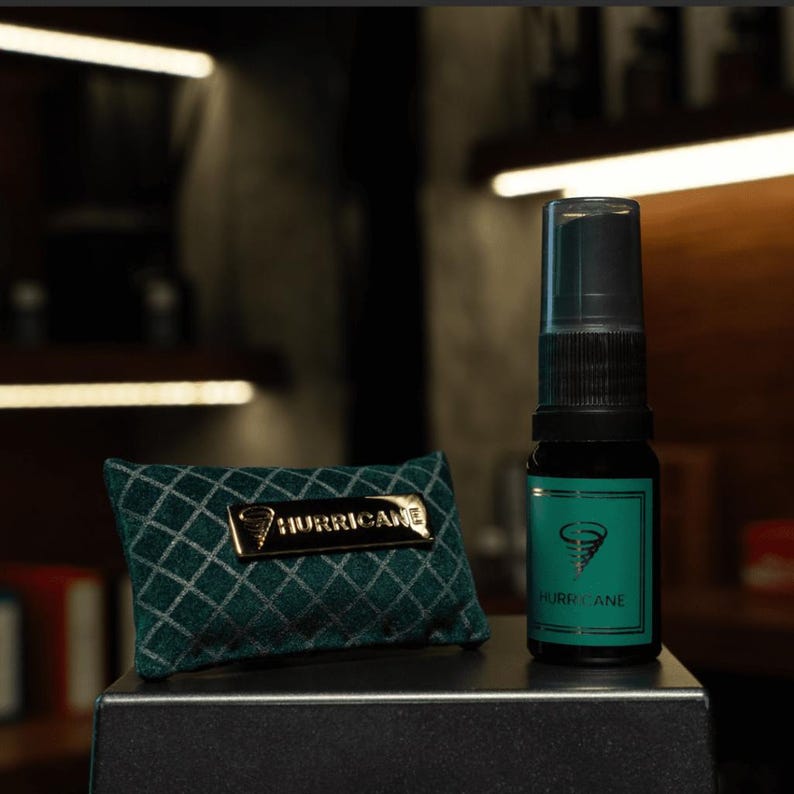 May include: A black spray bottle with a teal label and the word "HURRICANE" is next to a small, green pillow with a gold "HURRICANE" emblem. The bottle has a black cap and the pillow has a diamond pattern.