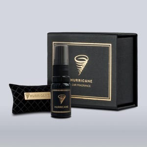 May include: A black car fragrance set with the word "HURRICANE" in gold lettering. The set includes a spray bottle, a small pillow, and a box, all in black. The bottle and box feature a gold tornado graphic.