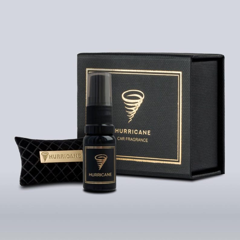May include: A black car fragrance set with the word "HURRICANE" in gold lettering. The set includes a spray bottle, a small pillow, and a box, all in black. The bottle and box feature a gold tornado graphic.