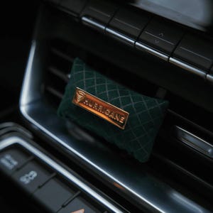 May include: A dark green, diamond-patterned car air freshener with a gold-colored rectangular plate that reads "HURRICANE". The air freshener is rectangular and fits into a car's air vent.