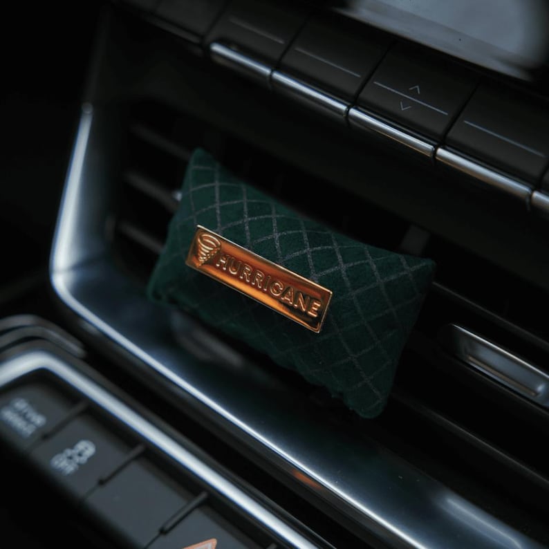 May include: A dark green, diamond-patterned car air freshener with a gold-colored rectangular plate that reads "HURRICANE". The air freshener is rectangular and fits into a car's air vent.