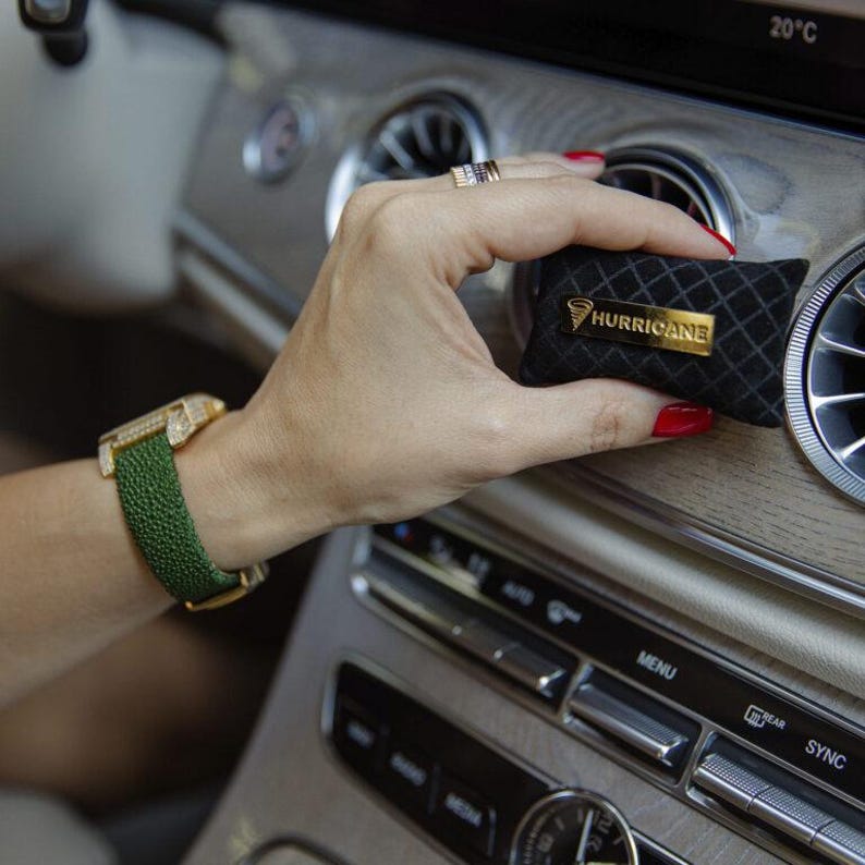May include: A black car air freshener with a gold "HURRICANE" logo is held near a car vent. The air freshener has a diamond pattern. A green watch with a gold case is on a wrist.