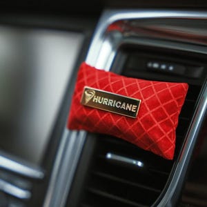 May include: A red, diamond-patterned air freshener with a gold-colored rectangular label that reads "HURRICANE". The air freshener is attached to a car's air vent, with a blurred background.